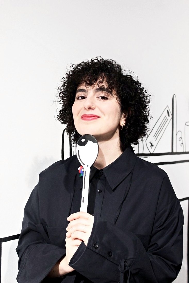 Ada Güvenir holding a painted spoon in front of one of her wall drawings